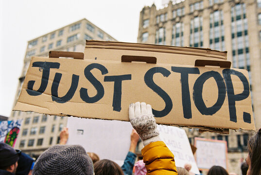 Protester holds cardboard sign saying "JUST STOP" at a political rally