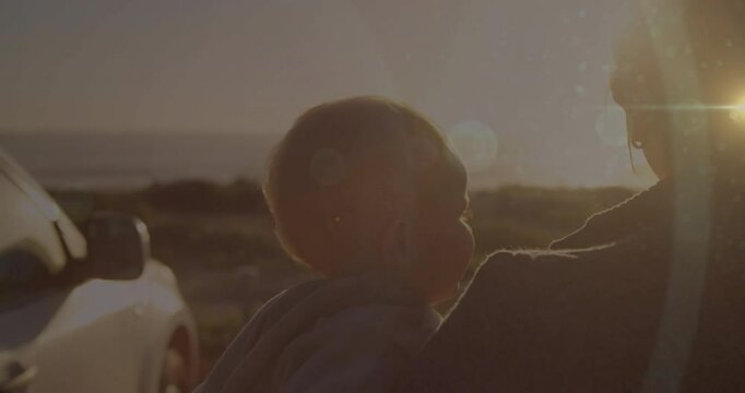 Mother stepping from car beside sea, cradling baby, gazing sunset with lens flares for health