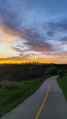 Paved path with a yellow line curving towards Calgary's downtown skyline under a dramatic sunset sky with orange and blue clouds