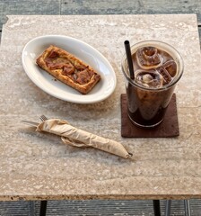 Top view of iced coffee and a slice of pizza on a marble table, served with wrapped cutlery in a cozy café setting
