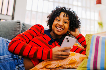 Woman relaxing on sofa and laughing while using smartphone