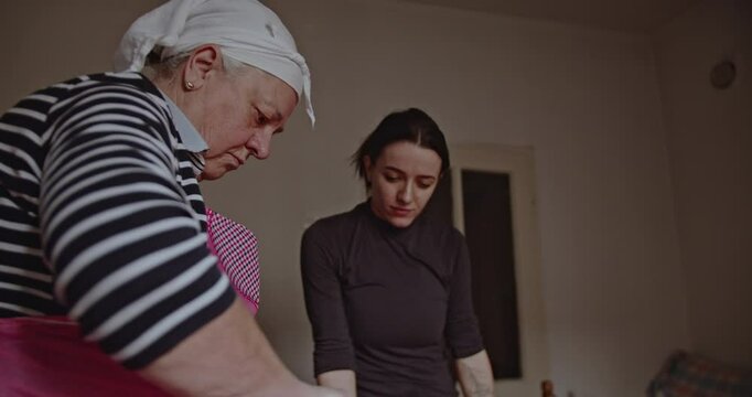 A senior woman wearing an apron and headscarf teaches a younger woman how to make a traditional dish in a cozy kitchen.