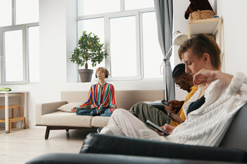 young adult woman meditating while colleagues and friends use smartphones in modern workplace,...