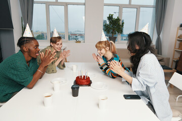 Young adult colleagues celebrating birthday with cake at workplace, wearing party hats and...