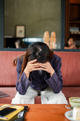 A woman in a blazer sits on a couch with her head in her hands, conveying stress or contemplation...