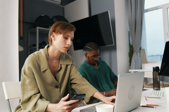 focused woman working on laptop and phone with coworker in modern workplace, young adult colleagues socializing, Gen Z business environment