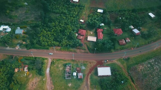 Overhead tracking drone shot of a white car driving through a rural road lined with banana groves, red-roofed homes, and scattered buildings in the Mount Elgon region of Eastern Uganda.