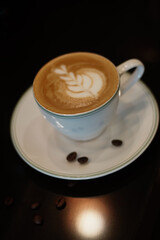 A white ceramic cup sits on a matching saucer, containing a latte with intricate froth art resembling a leaf pattern
