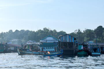 A vibrant scene unfolds with a colorful fishing boat navigating calm blue waters past a stilted village featuring weathered wooden structures and lush green trees under a clear sky