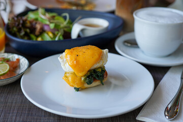 Delicious oozing eggs florentine on white plate with a cup of hot cappuccino, salad dish background
