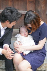 In June 2025, a young Japanese couple in their early twenties sits on a stone wall at a shrine in Aichi, soothing their one-month-old baby with a traditional den-den daiko during omiyamairi.