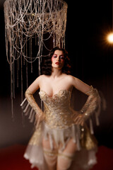 Elegant young circus actress in golden corset and gloves, standing under chandelier, front view....