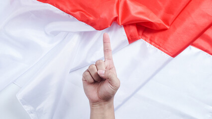A hand making a gesture with the index finger pointing upward symbolizes number one or unity in front of the red and white Indonesian flag to celebrate Indonesia independence day