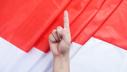 A hand making a gesture with the index finger pointing upward symbolizes number one or unity in front of the red and white Indonesian flag to celebrate Indonesia independence day
