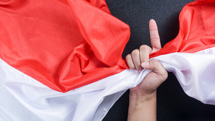 A hand holding red and white flag of Indonesia while making a gesture symbolizes number one or unity to celebrate Indonesia independence day isolated on black background