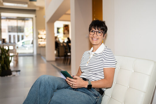 Optician smiling and using smartphone while sitting in optical store