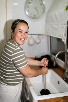 Woman unclogging kitchen sink with plunger and smiling at camera