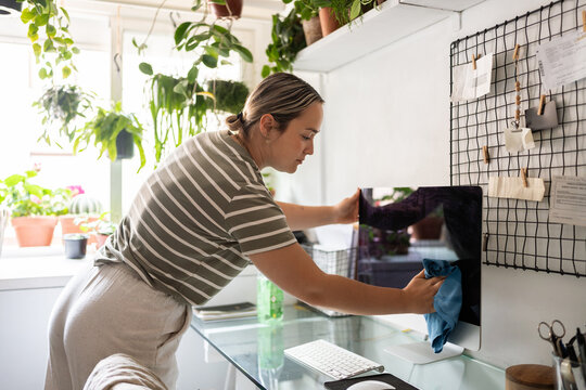 Woman cleaning computer screen in her home office surrounded by plants