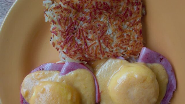 Zoom out from crispy hash browns with melted cheese, ham and eggs on plate.