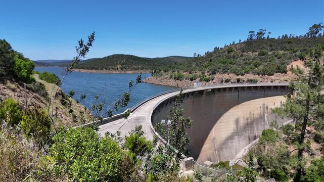 Bravura Dam curving along Odeleite River with surrounding hills and vegetation under a clear blue sky in Portugal
