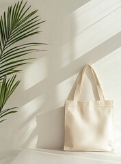 A cream-colored canvas tote bag hangs from a white wall, illuminated by sunlight casting shadows.  A palm leaf plant adds a touch of nature to the scene