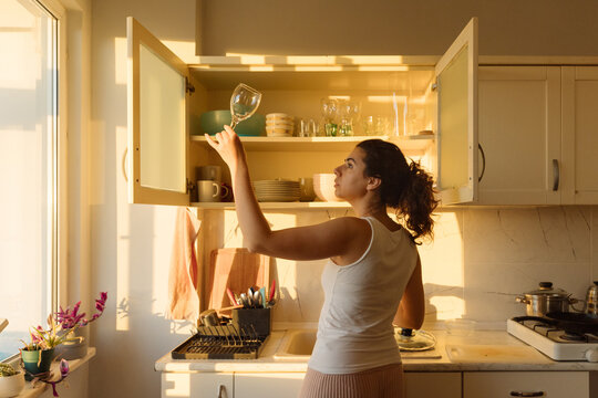 A young woman places glasses in a kitchen cabinet.