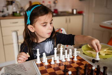 Little girl playing chess and eating apple slices at home