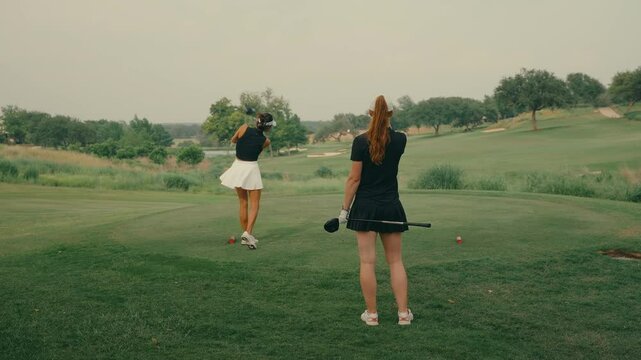 Female golfer hits a driver off the tee box in slow motion while her playing partner watches from behind. Captures focus, athleticism, and camaraderie on a sunny outdoor golf course.