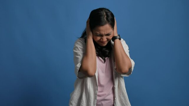 Unhappy sad angry young asian woman close eyes and cover ears by hands being annoyed with loud noise, isolated blue studio background, emotions, dislike loud sounds