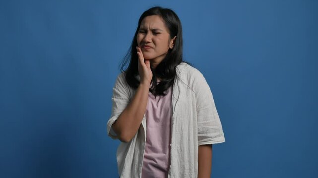 Unhappy young Asian woman touch cheek, suffers from toothache, having terrible sudden tooth pain isolated on blue background