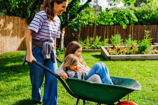 Mother pushing happy daughters in wheelbarrow in backyard garden