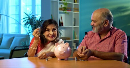 Indian senior couple promoting saving money investment habits for future security, mature husband wife placing coins in piggy bank at home, planning retirement, budget, financial awareness concept