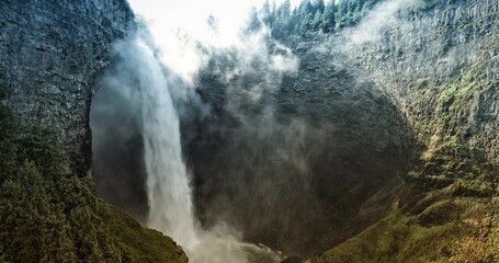 Narrow waterfall is plunging down rugged cliff in flat design, with mist, moss, evergreen trees