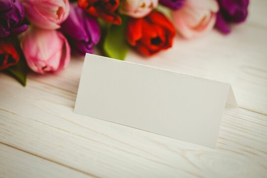 Fototapeta folded white place card is sitting on whitewashed tabletop, showing colorful tulip bouquet backdrop