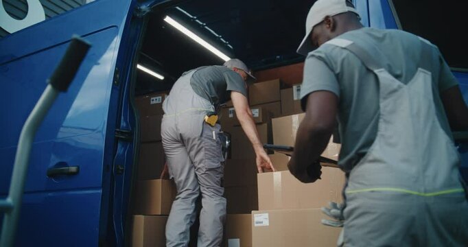 Outside of Logistics Warehouse: Male Employee Unloading Delivery Truck with Cardboard Boxes, Online Orders, Purchases and E-Commerce Goods. African American Manager Using Digital Tablet. Slow Motion. - Powered by Adobe