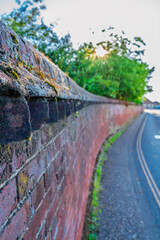 Sunlit country lane with historic stone wall featuring moss and lichen growth. Traditional stonework boundary along a quiet rural road with natural lighting