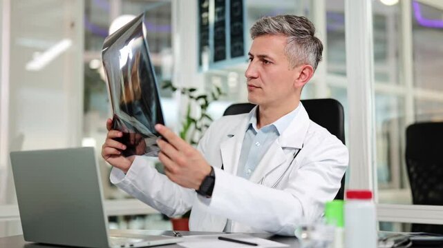 Professional male doctor in a white lab coat and stethoscope carefully examines an X-ray scan in a modern clinic office. He appears focused on patient diagnosis and health. - Powered by Adobe