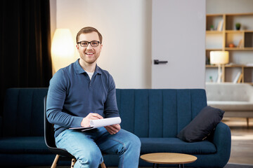 Fototapeta premium Portrait of happy male psychologist looking at camera and taking notes during therapy session at clinic. Friendly psychotherapist posing and smiling at office
