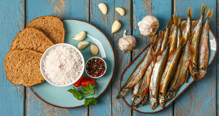 Plates with tasty smoked capelin, bread pieces, garlic and sea salt on blue wooden background