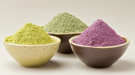 Three bowls of powdered food coloring.  Pale yellow, muted green, and deep purple powders are piled high in shallow, ceramic bowls.  A plain, light beige background isolates the bowls