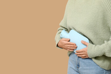 Young woman with hot water bottle having menstrual cramps on beige background, closeup