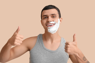 Handsome young man with shaving foam on his face showing thumbs-up gesture against beige background