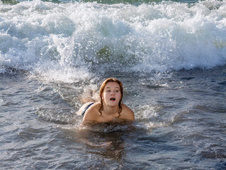 A young girl lying in the water on the shore opened her mouth in surprise and fear because of the foaming wave rolling over her from behind