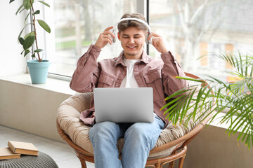 Male student with laptop putting on headphones before studying online in armchair at home