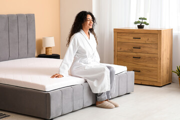Young African-American woman sitting on soft mattress in bedroom
