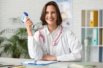Beautiful female doctor with infrared thermometer and stethoscope at table in clinic