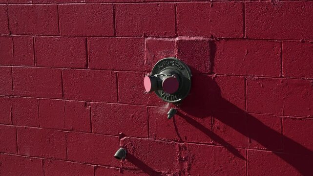 Close-up view of a fire department connection mounted on a vivid red brick wall. Harsh sunlight creates a long shadow, highlighting the industrial texture and minimal urban aesthetic.