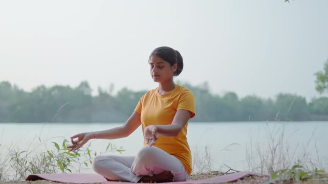 indian woman sitting in lotus position changing gyana mudra to anjali mudra by a serene lake, morning sunlight falling on her face.