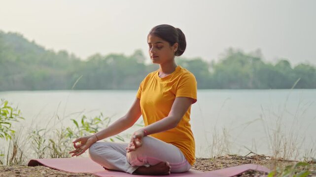 Attractive indian woman meditating with gyana mudra by a serene lake, morning sunlight falling on her face.