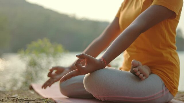 Tight shot of a woman sitting in padmasana pose on a yoga mat during sunrise at outdoor.